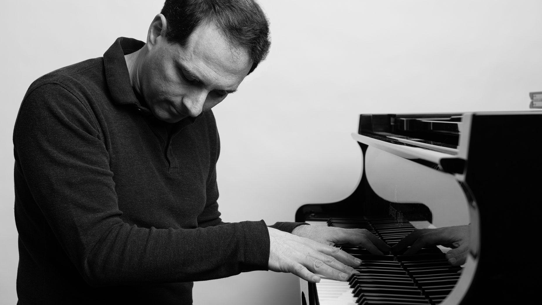 Alex Heffes, composer, arms crossed against a grey backdrop beside his name in bold type.
